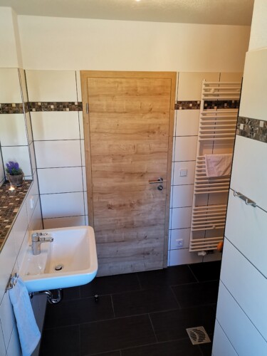 Modernes Badezimmer mit Holztür, weiß gefliesten Wänden, Wandwaschbecken, Handtuchhalter und einer kleinen Topfpflanze. Der saubere, helle Raum ist typisch für Exklusive Ferienhäuser Sächsische Schweiz - ideal für Ihren Wanderurlaub Sächsische Schweiz.