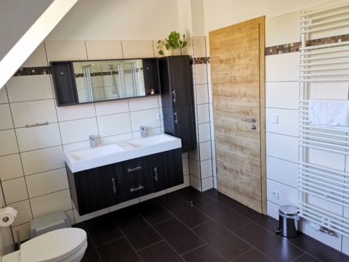 Modernes Bad in den exklusiven Ferienhäusern Sächsische Schweiz mit Doppelwaschbecken, dunklen Schränken, großem Spiegel, weißen Fliesen, Holztür, Handtuchheizung, Toilette und einem Pflanzenarrangement auf dem Schrank.