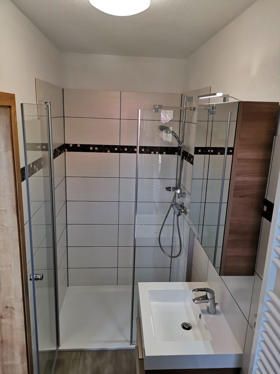 Modernes Bad in der Sachsenstiege mit Glasduschabtrennung, weißem Waschbecken mit verchromter Armatur, wandmontierter Duschbrause und hellen Fliesen mit einem dunklen Zierstreifen. Der Raum wirkt sauber und minimalistisch.
