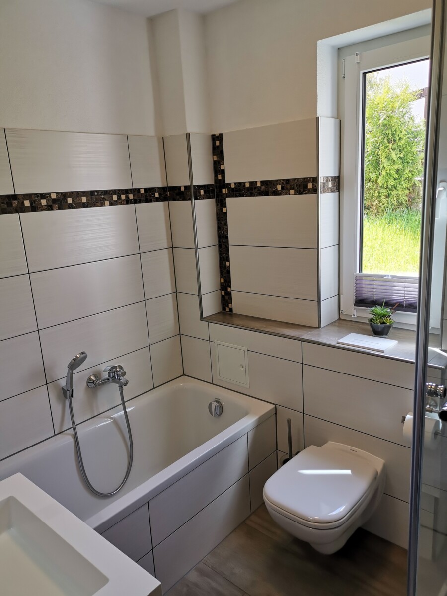 Modernes Wohnungsbad mit Badewanne, wandmontierter Toilette und weißen rechteckigen Fliesen, die durch eine dekorative schwarz-braune Bordüre akzentuiert werden. Natürliches Licht fällt durch ein Fenster an der Sachsenstiege in den Raum, wo eine kleine Pflanze auf dem Fensterbrett steht.