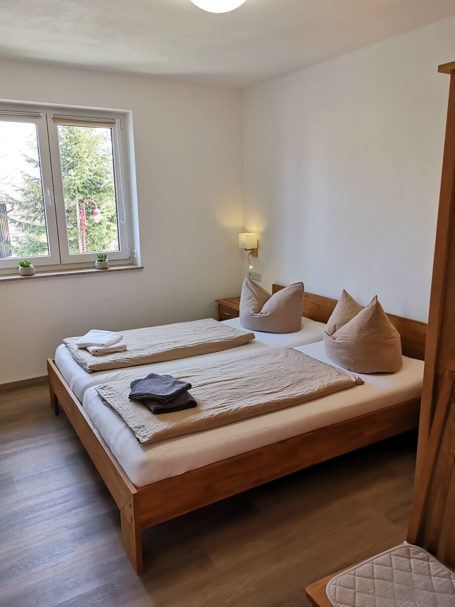 Ein gemütliches Schlafzimmer in der Sachsenstiege mit einem Doppelbett, zwei beigen Kissen, gefalteten Decken und Handtüchern. Das Zimmer ist mit Holzmöbeln, einem Fenster mit Topfpflanzen und sanftem Tageslicht ausgestattet.