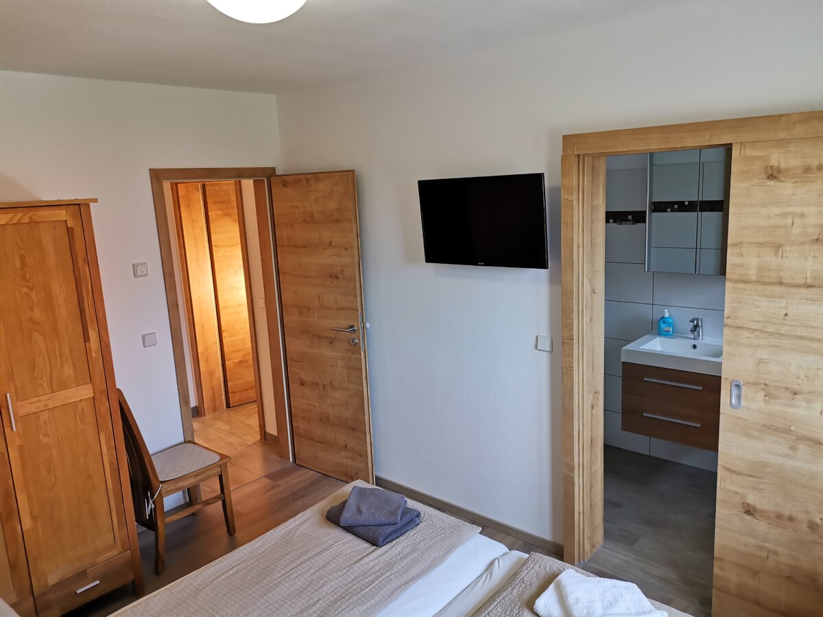 Ein gemütliches Schlafzimmer im Apartment Sachsenstiege ist mit Holzmöbeln, einem Kleiderschrank und einem Stuhl ausgestattet. Im Vordergrund steht ein Bett mit gefalteten Handtüchern. An der Wand befindet sich ein Fernseher und eine offene Tür zum Badezimmer mit Waschbecken.