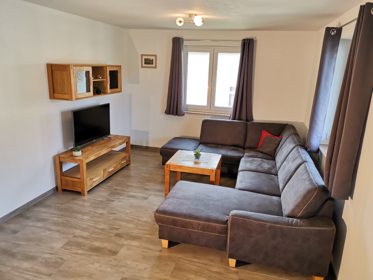 Das Wohnzimmer einer gemütlichen Wohnung an der Heiligen Stiege besteht aus einem großen grauen Sektionssofa, einem Couchtisch aus Holz, einem Fernseher auf einem Holzständer, Wandregalen, zwei Fenstern mit grauen Vorhängen, hellen Wänden und Holzböden.