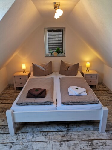Ein gemütliches Dachgeschoss-Schlafzimmer mit Dachschrägen im Exklusive Ferienhäuser Papstdorf, ausgestattet mit einem weißen Doppelbett mit braunem Bettzeug, Nachttischen mit Lampen, gefalteten Handtüchern und einem kleinen Fenster - perfekt für Ihren Wanderurlaub Sächsische Schweiz.