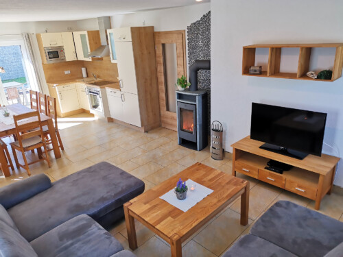 Modernes, offenes Wohnzimmer und Küche mit Holzmöbeln, grauem Sofa, TV, Esstisch, Holzofen und großen Fenstern, die viel Licht hereinlassen - perfekt für einen Wanderurlaub in der Sächsischen Schweiz oder einen erholsamen Aufenthalt.