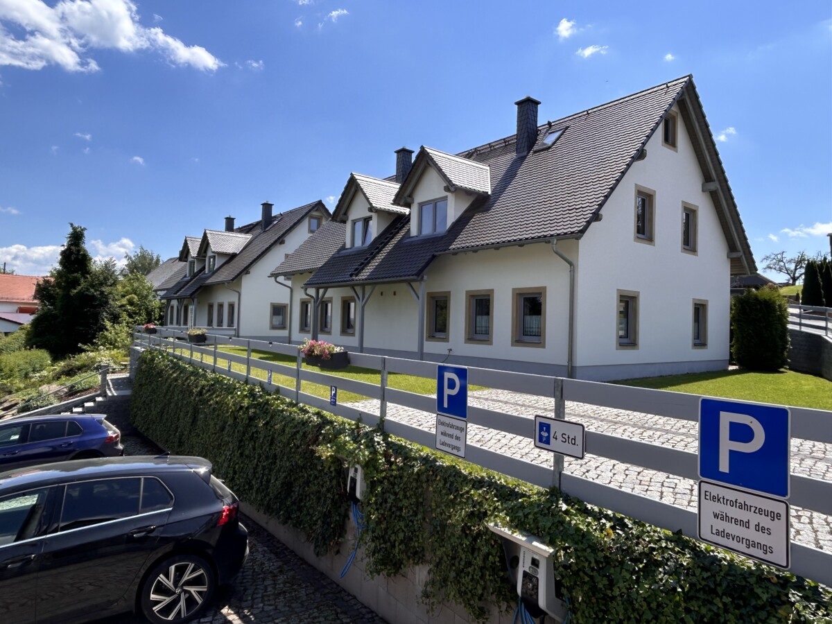 Eine Reihe moderner weißer Häuser mit dunklen Dächern steht hinter einem begrünten Zaun am Feriendomizil Vetter Papstdorf. Davor stehen mehrere geparkte Autos, blaue Parkschilder weisen auf Stellplätze für Elektrofahrzeuge unter meist klarem Himmel hin.