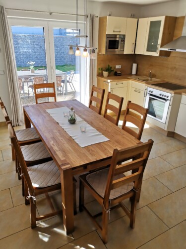 Heller, moderner Küchen- und Essbereich mit Holztisch für sechs Personen, weißen Schränken, Einbaugeräten und Glasschiebetüren, die auf eine sonnige Terrasse führen - perfekt für Ihren Wanderurlaub Sächsische Schweiz in Exklusive Ferienhäuser Papstdorf.