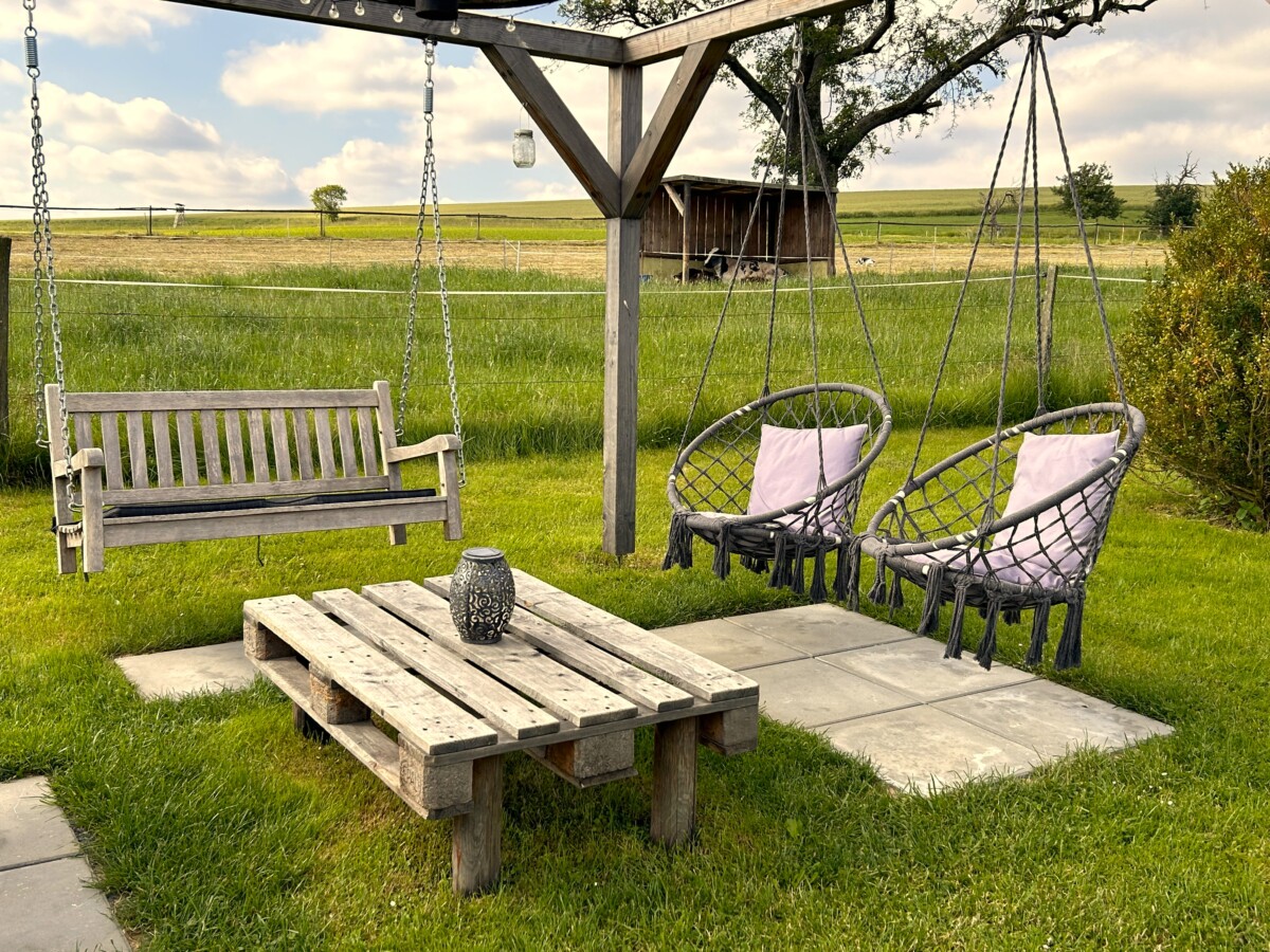Sitzplatz im Freien mit einer Holzschaukel, zwei Hängesesseln mit Kissen und einem Couchtisch aus Paletten auf einer Wiese in der Ferienanlage Vetter Gohrisch. Umgeben von Feldern, Bäumen und einem Holzschuppen im Hintergrund.