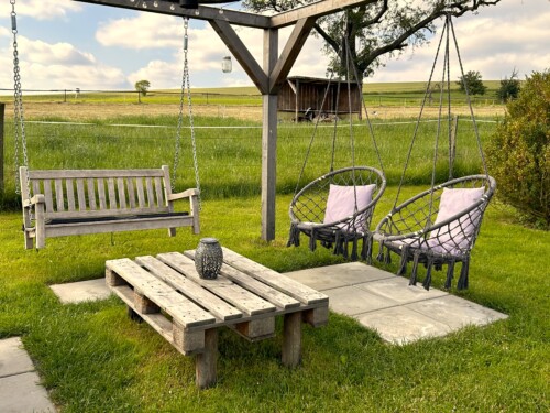 Die 5 Sterne Ferienhäuser Papstdorf verfügen über einen gemütlichen Sitzbereich im Freien mit einer Holzschaukel und zwei Hängesesseln unter einer Pergola. Genießen Sie die Aussicht auf offene Felder - perfekt für einen entspannten Wanderurlaub in der Sächsischen Schweiz.