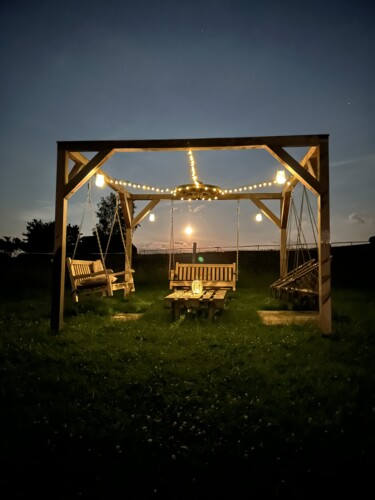 Eine Holzschaukel mit Bänken umgibt einen Tisch, der in der Abenddämmerung von Lichterketten beleuchtet wird. Der Mond geht über dem grasbewachsenen Rasen auf und schafft eine magische Szene im Feriendomizil Vetter Papstdorf im Herzen der Ferienanlage Sächsische Schweiz.