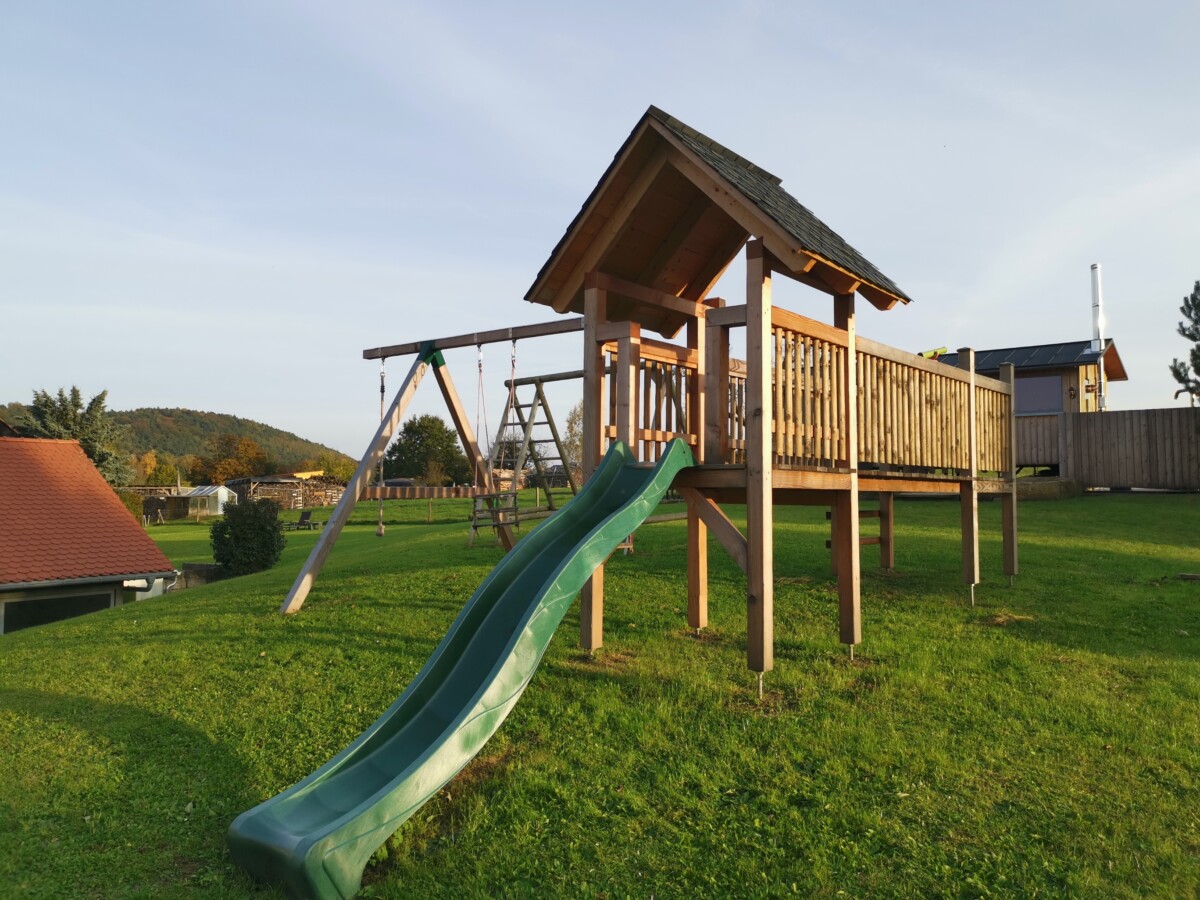 Ein hölzernes Spielgerät mit grüner Rutsche, Schaukeln und einem kleinen Dach steht auf dem grasbewachsenen Gelände des Feriendomizils Vetter Papstdorf, umgeben von Bäumen und Häusern unter einem klaren Himmel.