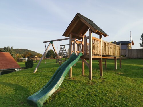 Ein hölzernes Spielgerät mit einer grünen Rutsche, einer Leiter, einer Schaukel und einem überdachten Podest steht auf einer Wiese bei den 5 Sterne Ferienhäusern Papstdorf, mit exklusiven Ferienhäusern Sächsische Schweiz und malerischen Bäumen im Hintergrund.
